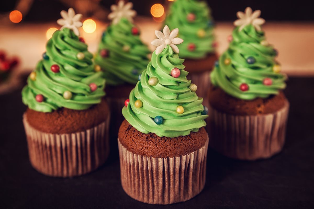 Un gruppo di cupcake natalizi decorati come piccoli alberi di Natale. Ogni cupcake ha una base di tortino marrone e una cima di glassa verde a spirale, ornata con palline di zucchero colorate e una stella di zucchero bianca in cima. Luci sfocate sullo sfondo.