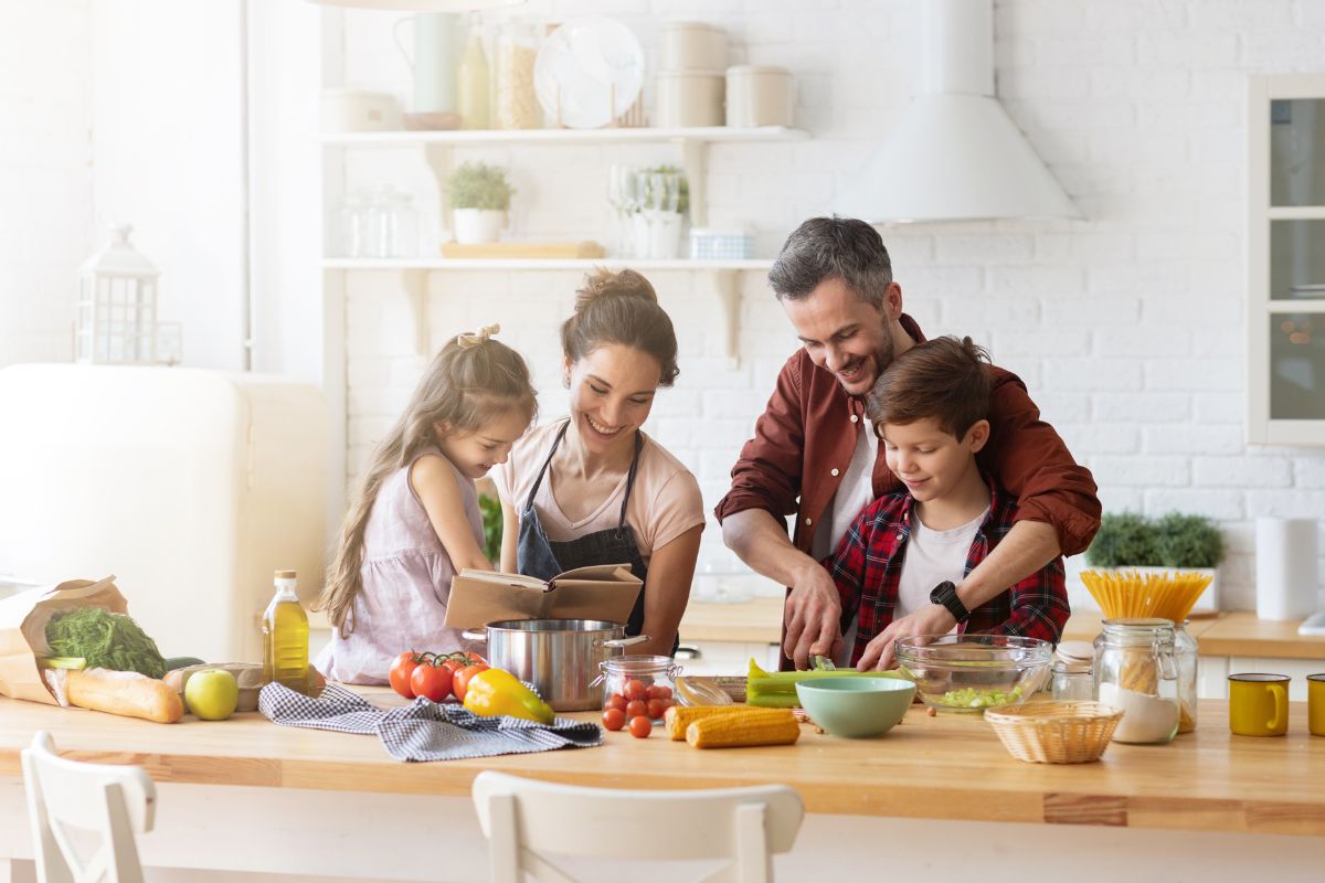 Famiglia (padre, madre, figlio e figlia) che cucina insieme in una cucina luminosa, preparando verdure e consultando un ricettario.