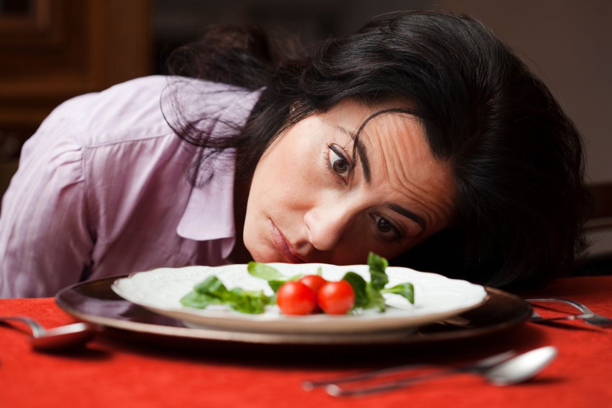Una donna con i capelli scuri, vestita con una camicia lilla, poggia la guancia su una tovaglia rossa, fissando con un'espressione di profonda insoddisfazione e tristezza un piatto bianco che contiene solo poche foglie di insalata verde e tre pomodorini rossi.