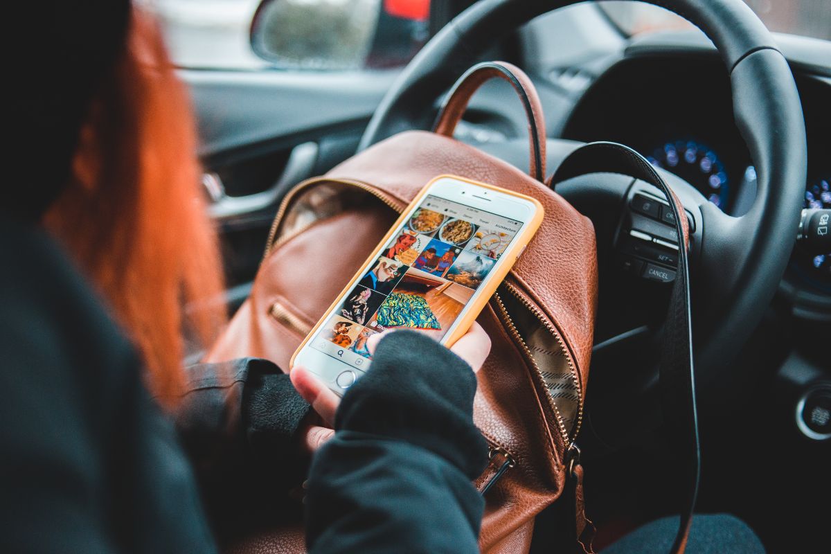 Una persona con capelli rossi e una felpa scura siede al volante di un'auto, tenendo in mano uno smartphone con custodia gialla che mostra un feed di immagini colorate, presumibilmente di cibo o opere d'arte. Uno zaino di pelle marrone chiaro è visibile sotto il telefono.