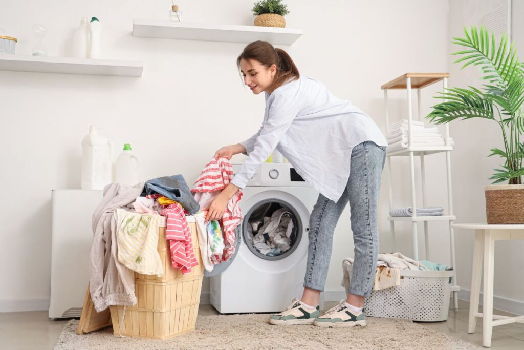 Una donna sorridente con una camicia azzurra e jeans si china per trasferire vestiti colorati da un cesto di vimini a una lavatrice bianca in una lavanderia luminosa.