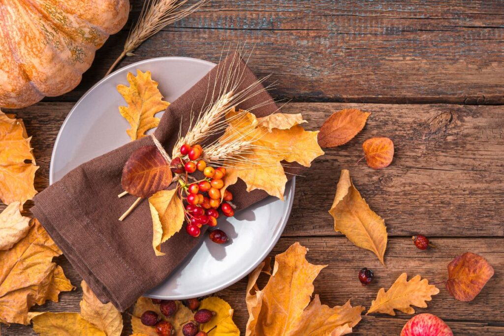 Centrotavola autunnale con piatto, tovagliolo marrone, foglie secche, spighe di grano e bacche arancioni, su un tavolo di legno scuro.