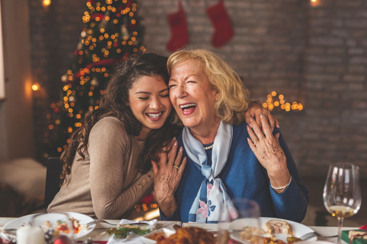Abbraccio e risate tra nonna e nipote (o due parenti) durante un pranzo di Natale, con albero e decorazioni sullo sfondo.