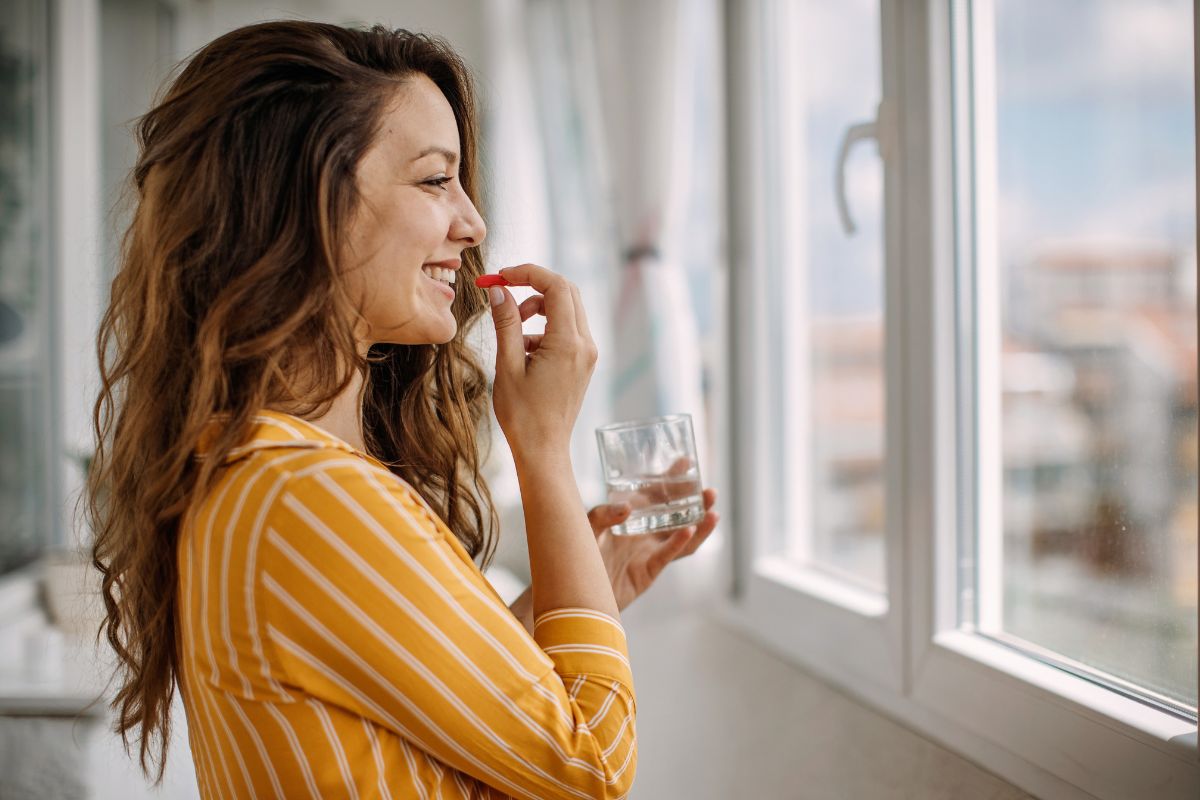 Donna sorridente in camicia gialla che prende una pillola rossa con un bicchiere d'acqua.