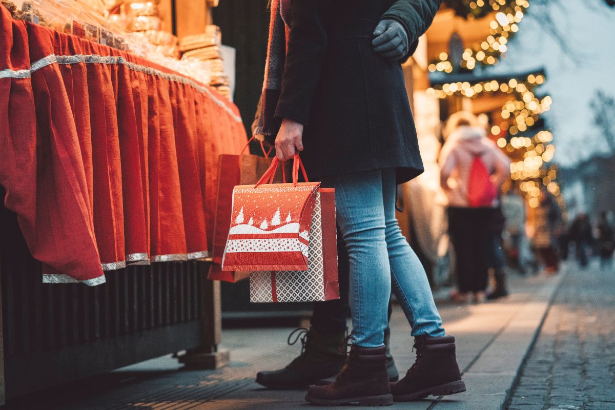 Coppia che tiene borse della spesa rosse tra le bancarelle illuminate di un mercatino di Natale.