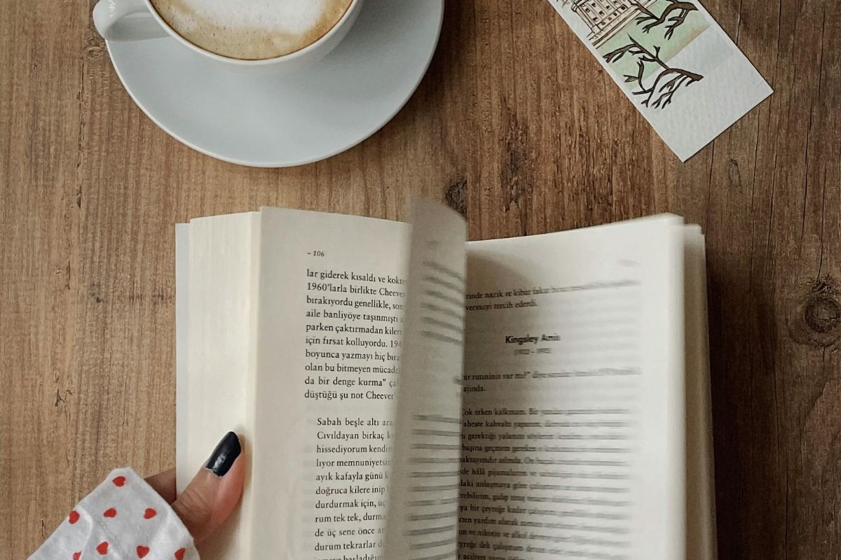 Mano che tiene un libro aperto con caffè e segnalibro su un tavolo di legno.