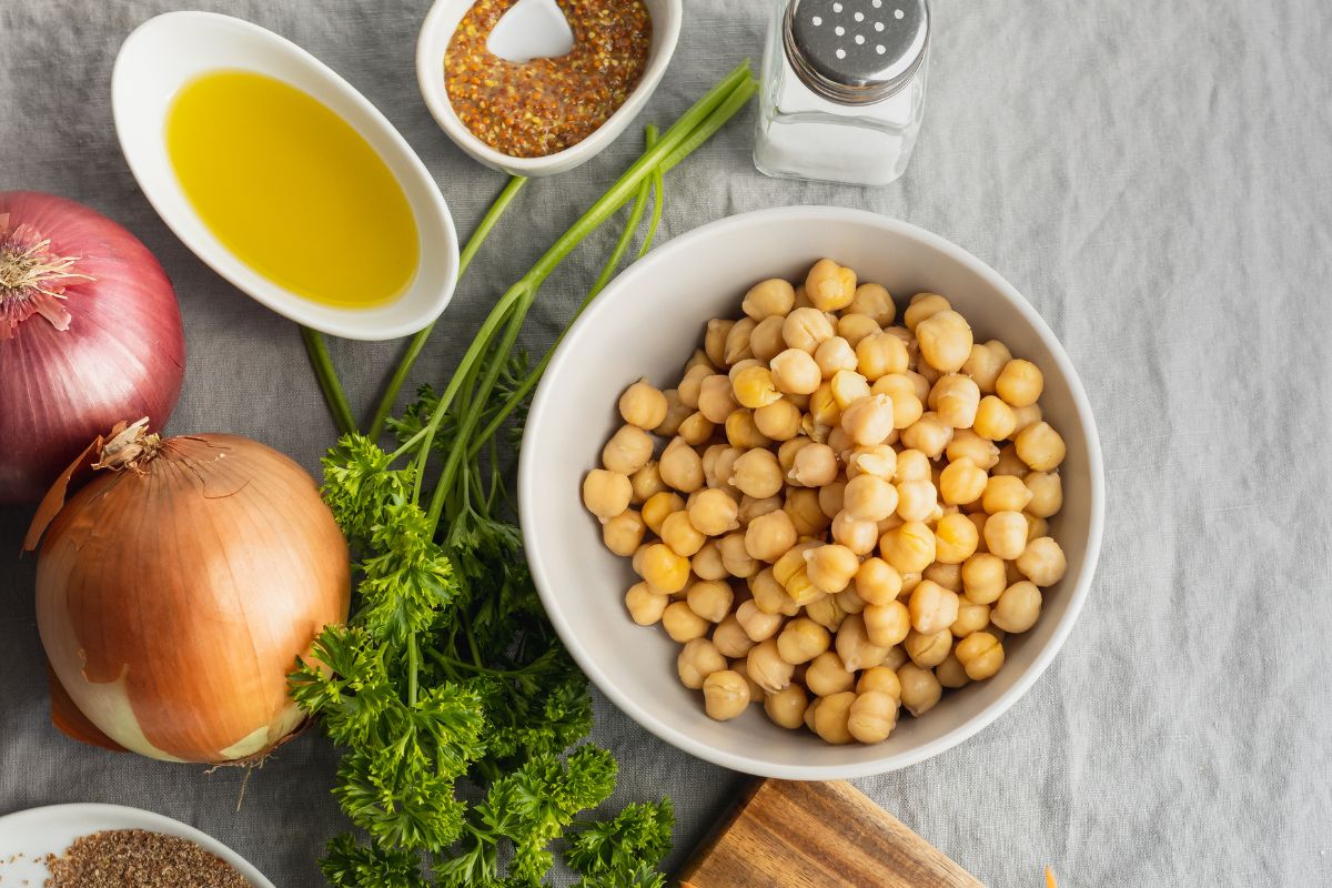 na vista dall'alto di ingredienti freschi su una tovaglia di lino grigio: una ciotola di ceci cotti, cipolle rosse e dorate, prezzemolo, una ciotola di olio d'oliva, senape in grani e una saliera.