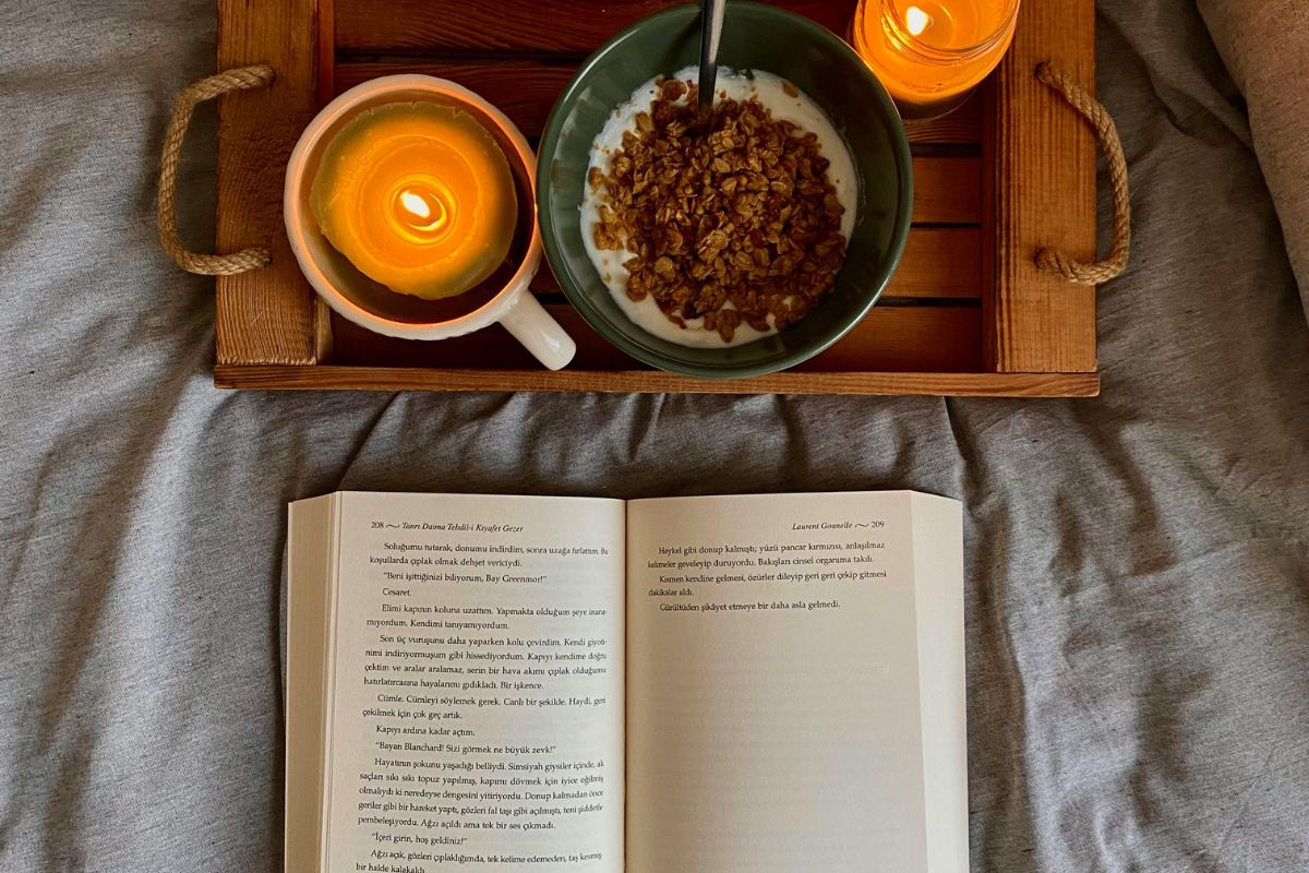 Un libro aperto su un letto accanto a un vassoio con colazione sana e candele accese.