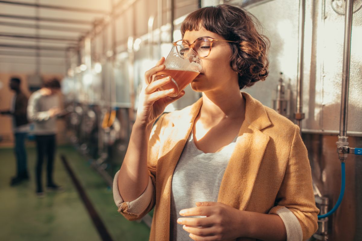Una giovane donna con i capelli corti e occhiali rotondi, vestita con una giacca color senape e una maglietta grigia, sta bevendo un sorso di birra chiara da un bicchiere in un ambiente industriale, probabilmente un birrificio, con serbatoi in acciaio inossidabile visibili sullo sfondo e luce solare che filtra da destra.