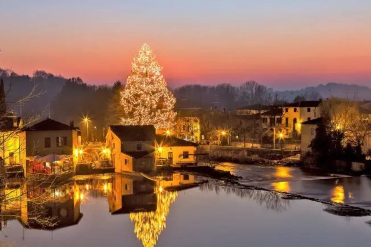 Paesaggio notturno di Borghetto sul Mincio con le luci natalizie accese, un grande albero illuminato e i riflessi sul fiume al tramonto.