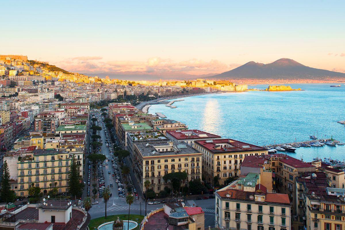 Veduta panoramica di Napoli al tramonto con il Vesuvio e il lungomare illuminato.