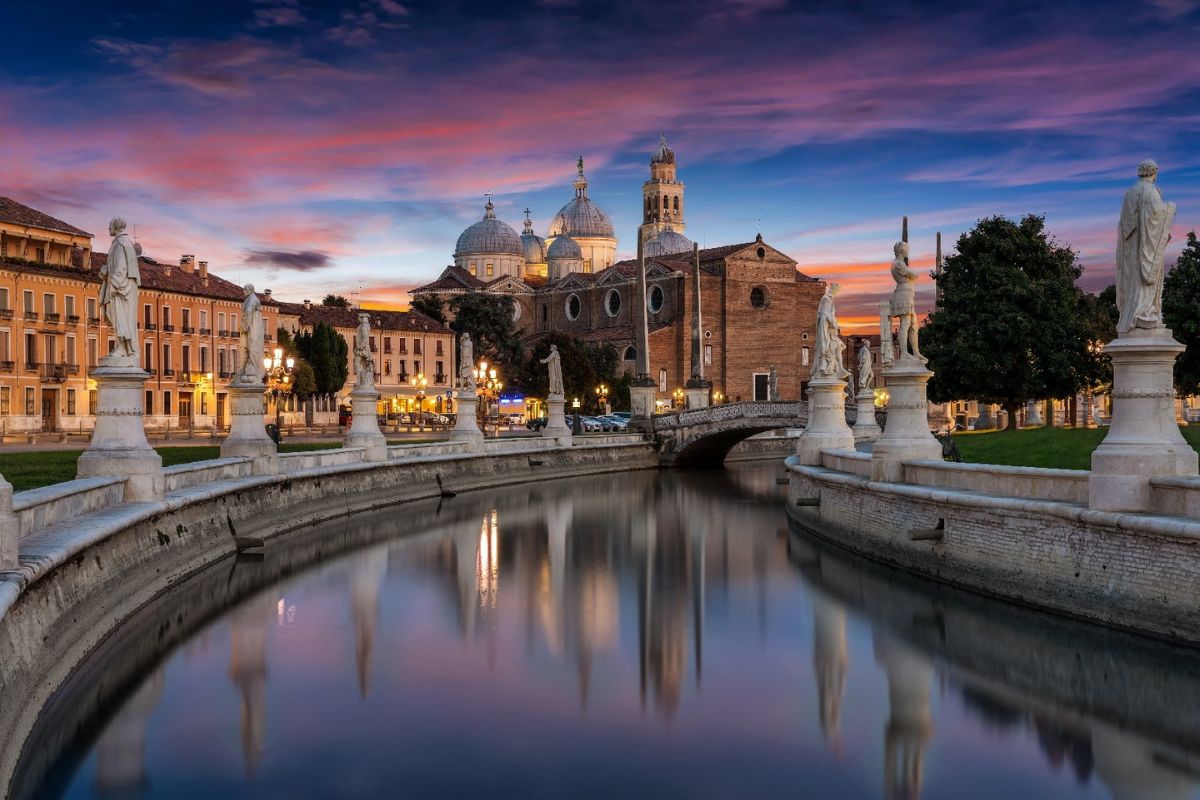 Vista panoramica del Prato della Valle a Padova al tramonto, con il canale, le statue e la Basilica di Sant'Antonio sullo sfondo.