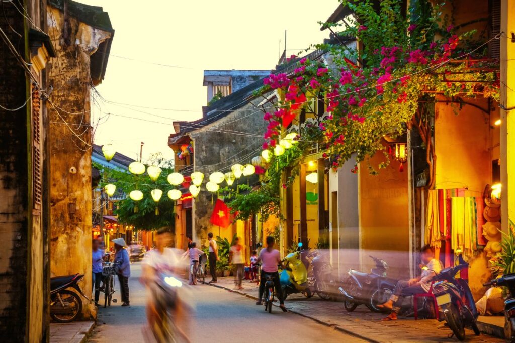 Una vivace strada notturna a Hoi An, Vietnam, illuminata da lanterne tradizionali appese sopra e da bougainvillea fiorita. Persone in movimento, moto parcheggiate e un mercante sono visibili.