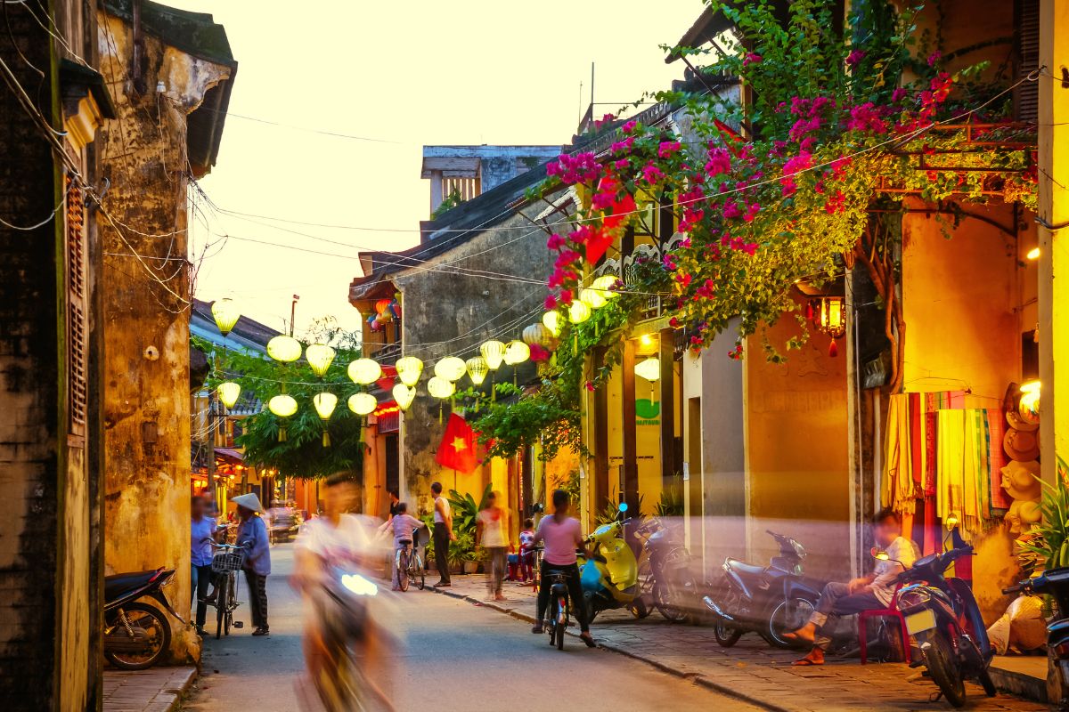 Una vivace strada notturna a Hoi An, Vietnam, illuminata da lanterne tradizionali appese sopra e da bougainvillea fiorita. Persone in movimento, moto parcheggiate e un mercante sono visibili.