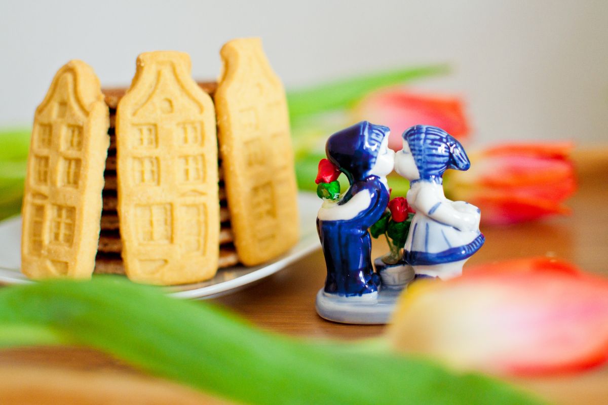 Composizione con biscotti a forma di case di Amsterdam e una statuina in ceramica Delfts Blue che raffigura due bambini che si baciano, su uno sfondo sfocato con tulipani.