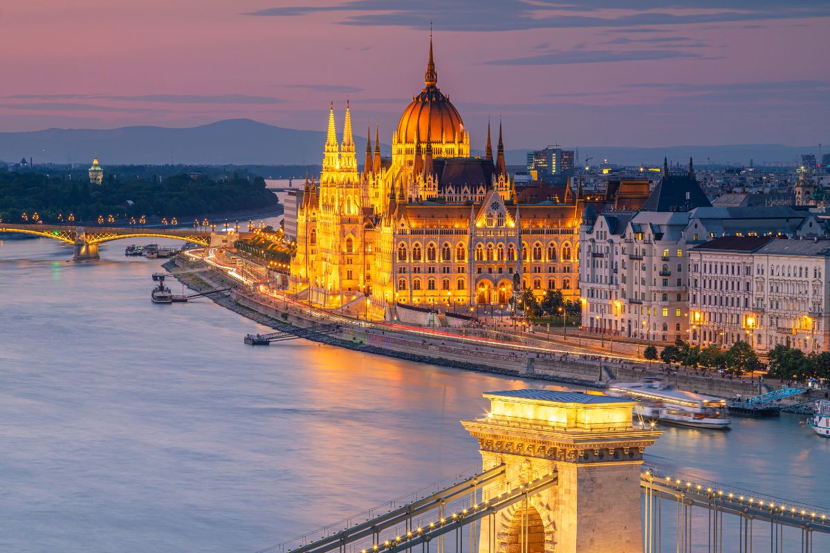 Veduta notturna e panoramica del Parlamento di Budapest illuminato sul fiume Danubio, con il Ponte delle Catene in primo piano.