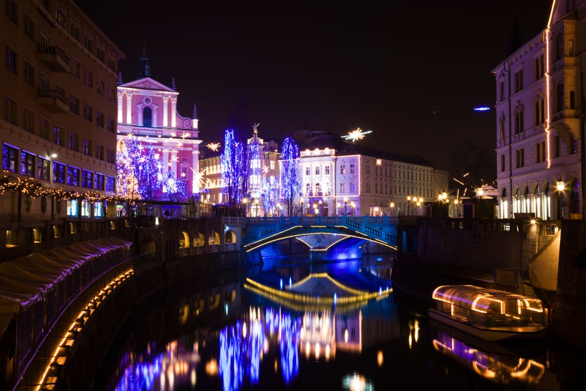 Un paesaggio urbano notturno di Lubiana, con un ponte illuminato di blu e edifici storici decorati con luci natalizie blu e rosa, riflettendosi sull'acqua scura.