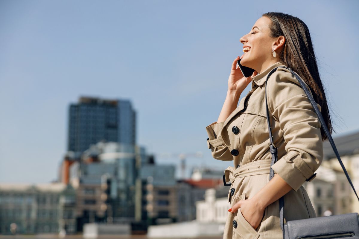 Donna sorridente al telefono con trench in ambiente urbano.