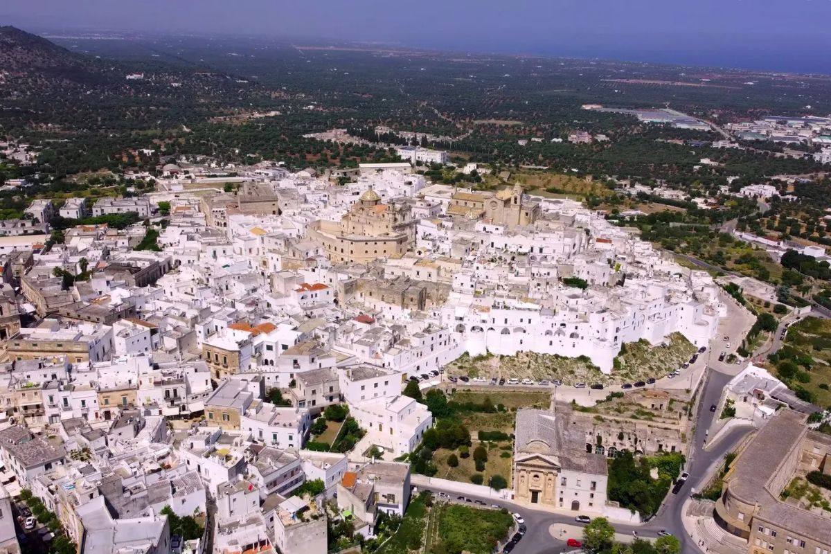 Veduta aerea della Città Bianca di Ostuni, Puglia, con case imbiancate che si arrampicano sulla collina verso la cattedrale e la campagna circostante piena di ulivi.
