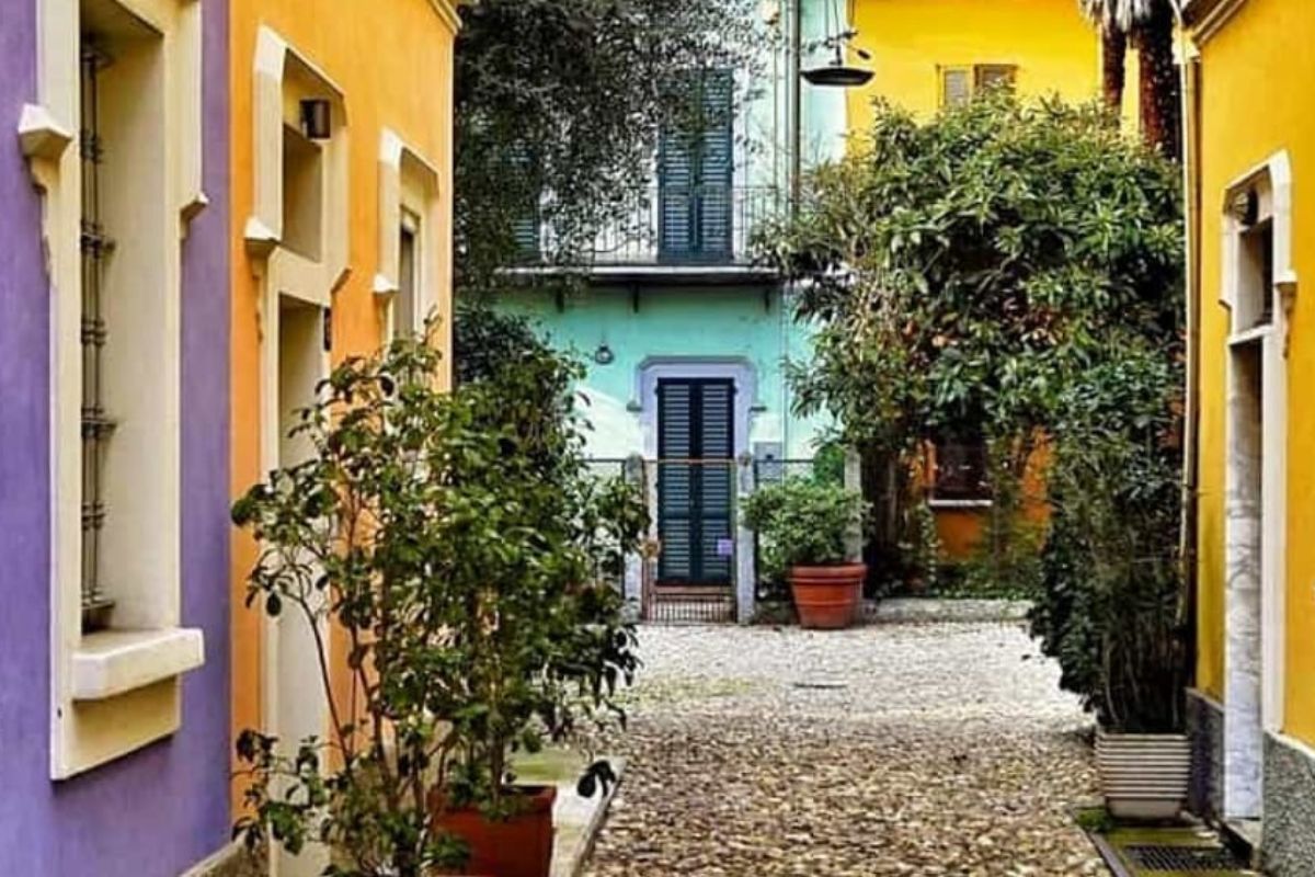 Vicolo lastricato e stretto di Via Abramo Lincoln a Milano, fiancheggiato da case dai colori vivaci (viola, arancio, giallo) e vegetazione.